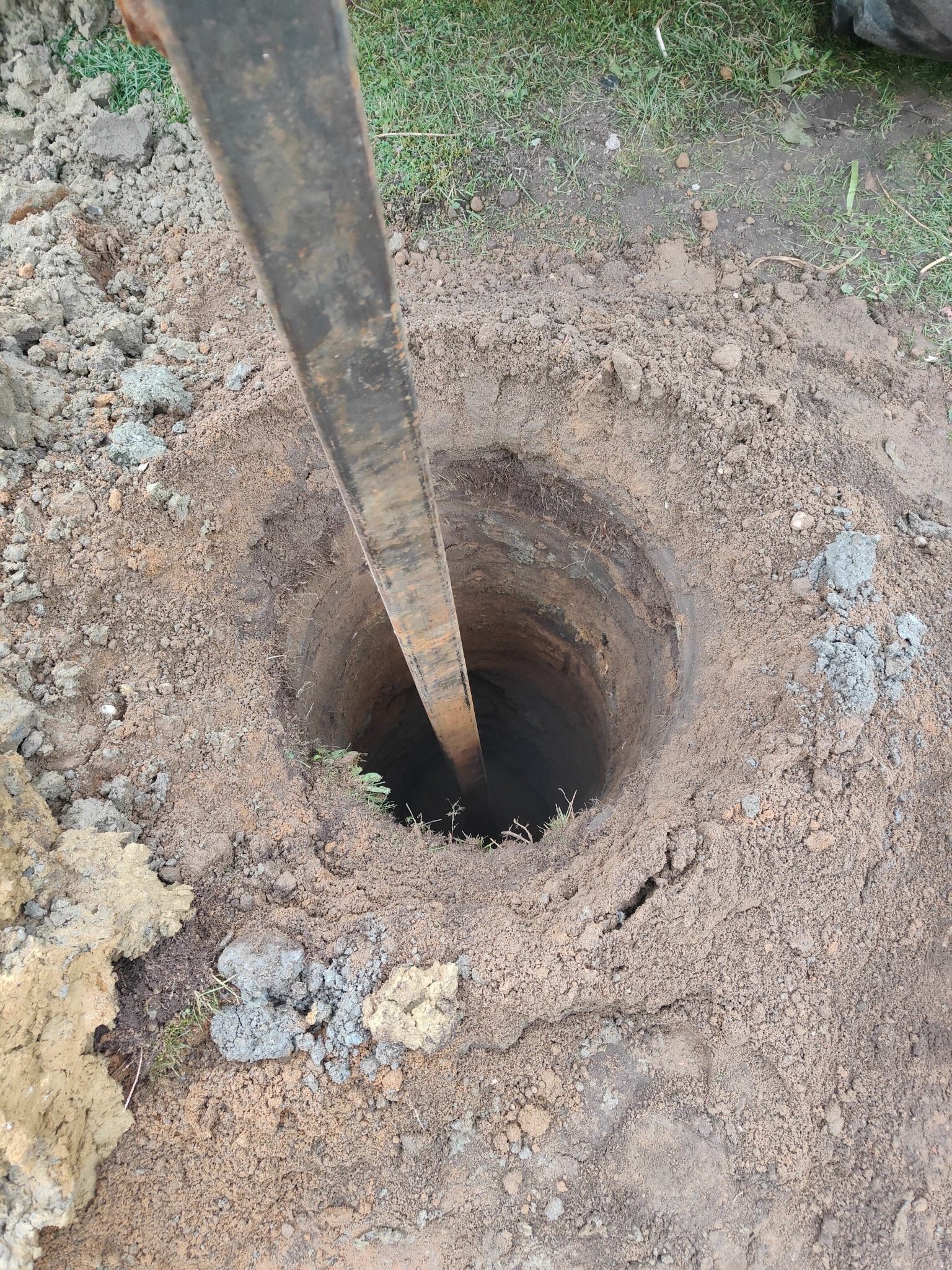 Wykopany okrągły otwór w ziemi z metalowym prętem wewnątrz, widoczna trawa na obrzeżach.