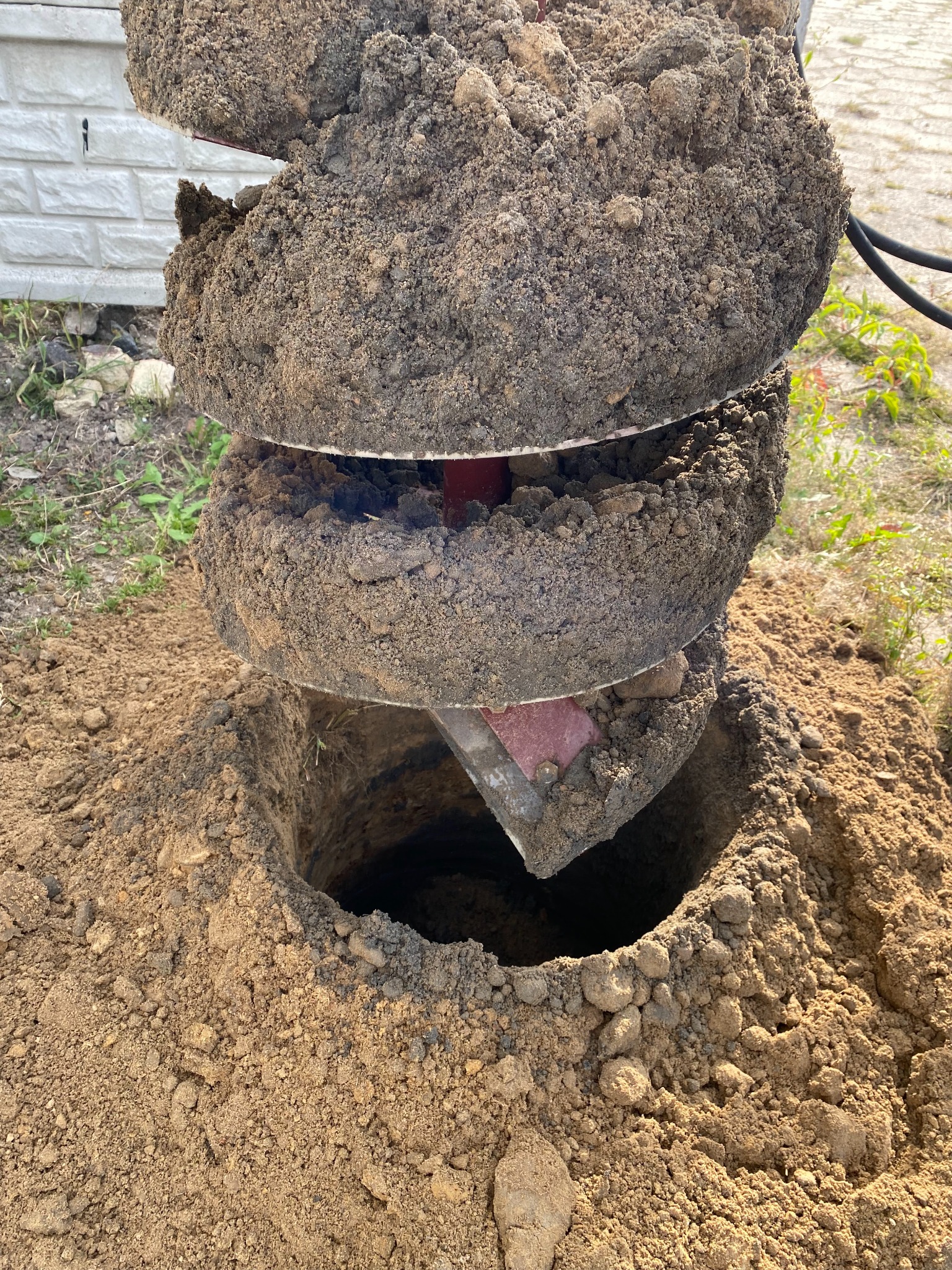 Wiertło świdrowe zagłębia się w ziemię, tworząc otwór pod instalację. Wokół otworu widoczny usypany grunt, a w tle fragment białego ogrodzenia.