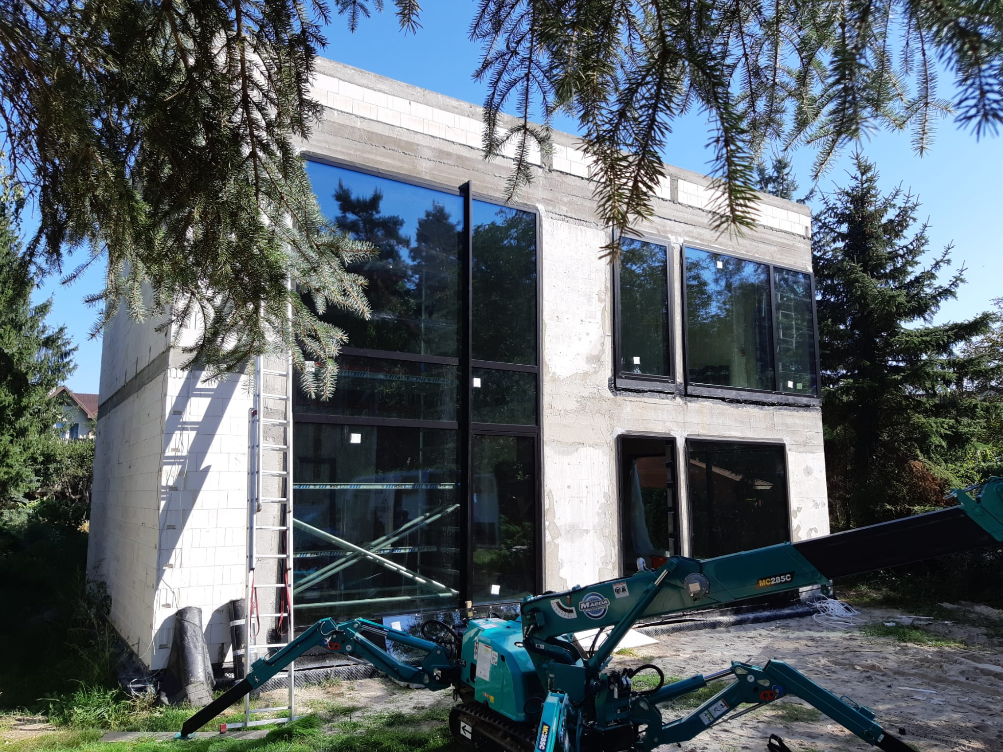 Surowy budynek z dużymi, czarnymi oknami, w trakcie montażu. Na pierwszym planie minikoparka, w tle drzewa i błękitne niebo. Widoczna drabina oparta o ścianę.