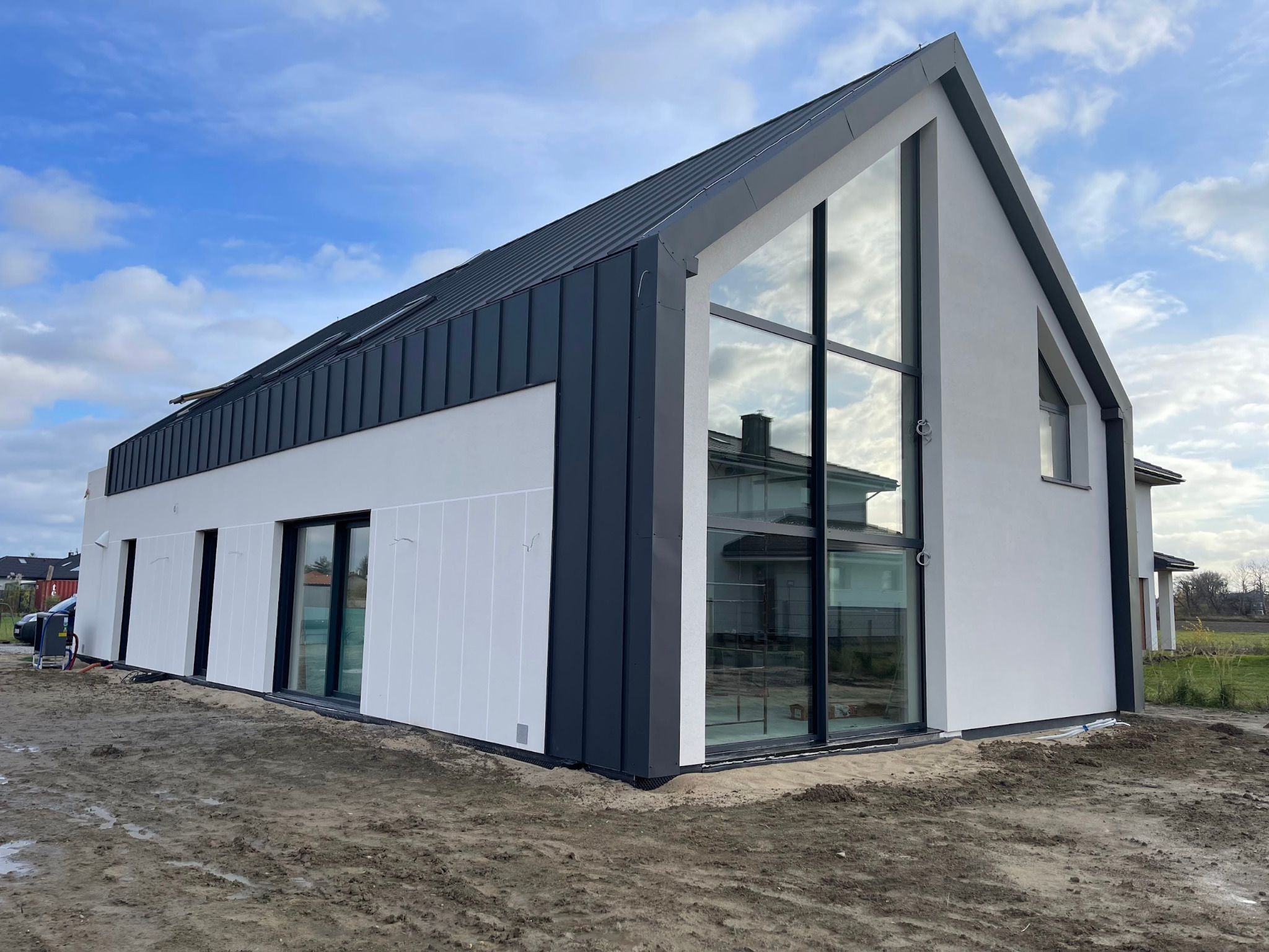 Nowoczesny dom z dużymi oknami, ciemną blachą na dachu i białymi ścianami, widziany z zewnątrz, w trakcie wykończenia terenu wokół budynku.