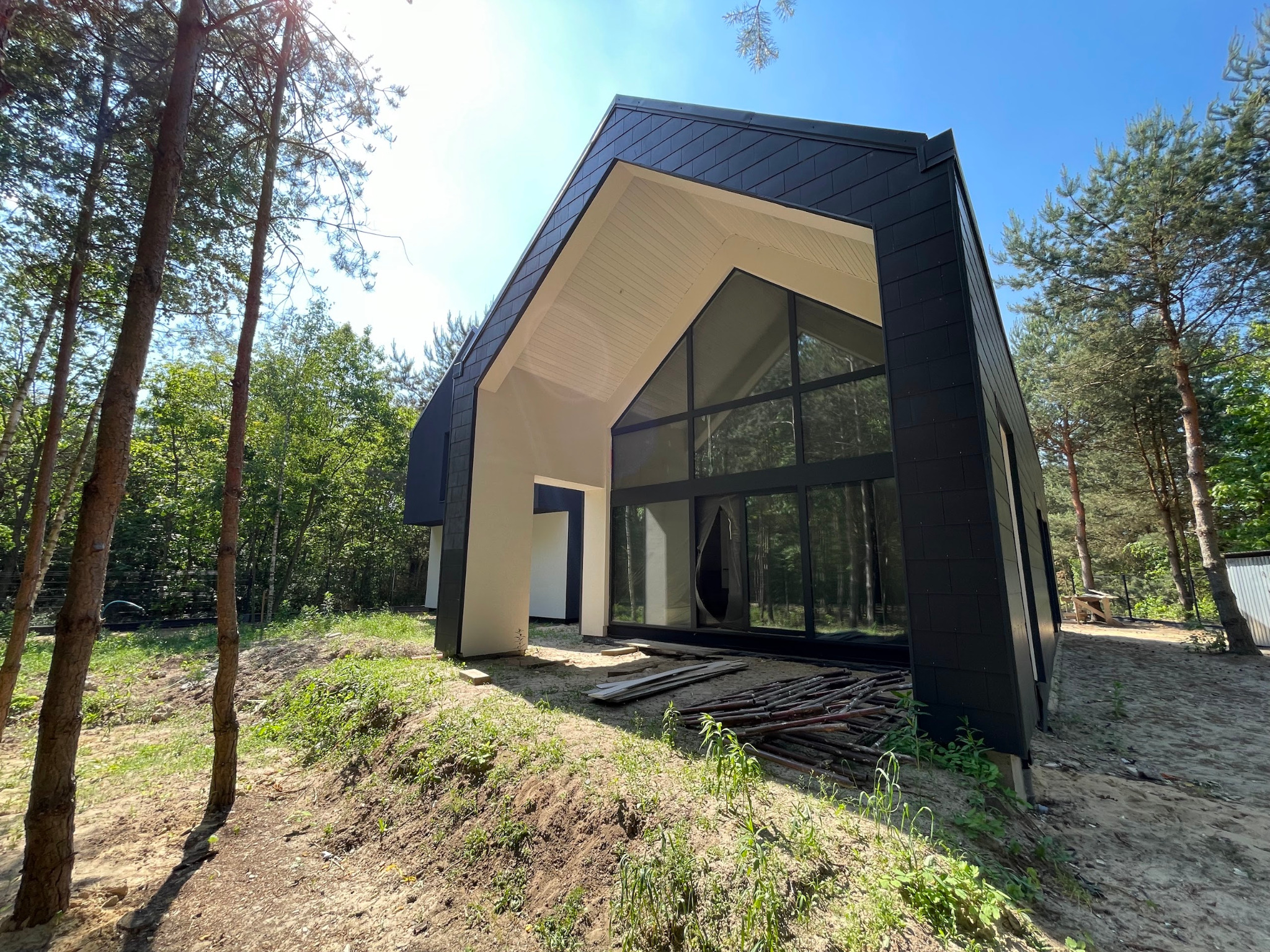 Nowoczesny dom z czarną elewacją i dużymi oknami, otoczony zielenią. Widoczne drewniane elementy i jasne wnętrze przez okna. Dom w stylu minimalistycznym.