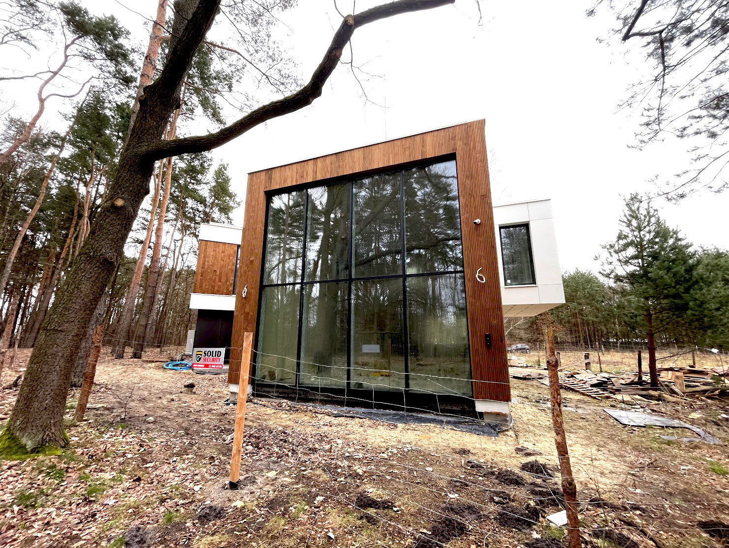 Nowoczesny dom z dużymi oknami w czarnych ramach, otoczony lasem. Widoczne drewniane elementy elewacji i numer domu '6'.