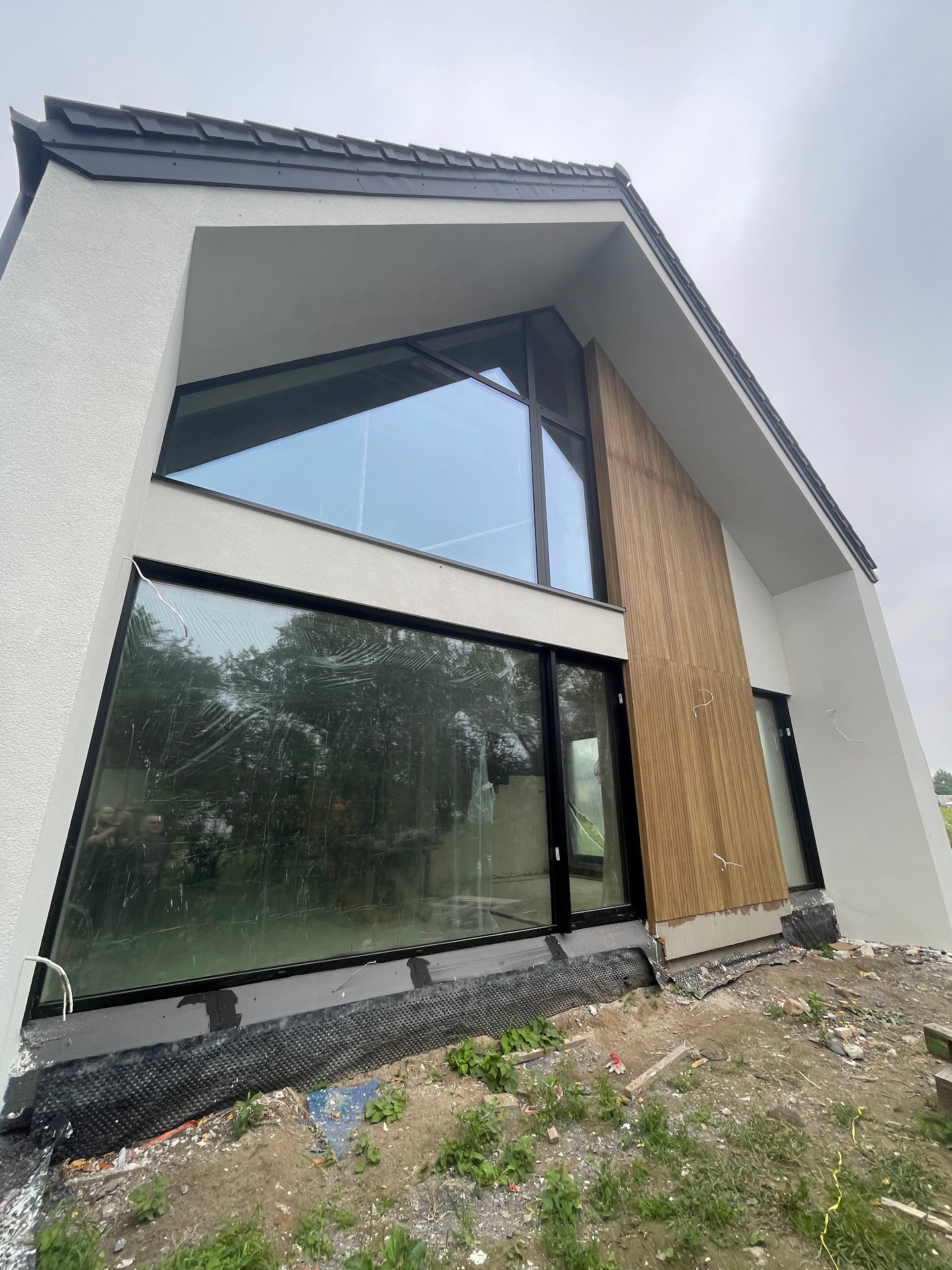 Nowoczesny dom z dużymi, trójkątnymi oknami w czarnych ramach i drewnianą fasadą. Widoczne folie ochronne na szybach. Surowy teren wokół budynku.