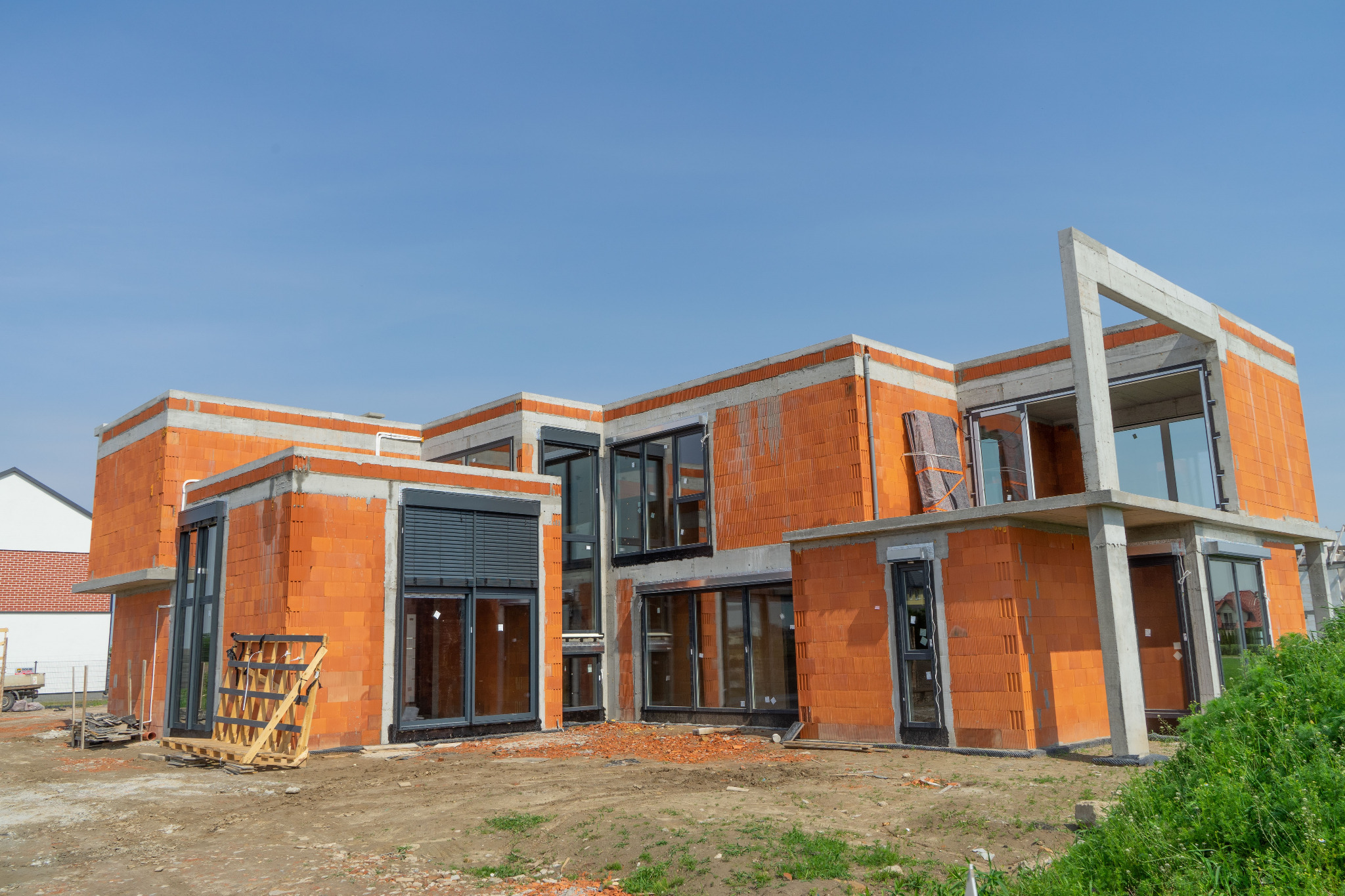 Budynek w trakcie budowy z czerwonej cegły, z zamontowanymi oknami i roletami zewnętrznymi. Widoczne elementy konstrukcyjne z betonu i drewniana paleta.