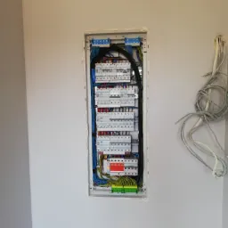 Wnęka w ścianie z rozdzielnicą elektryczną, widoczne liczne bezpieczniki, przewody w różnych kolorach (niebieski, czarny, zielony, żółty), po prawej stronie ściany luźne przewody wychodzące ze ściany.
