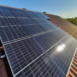 Panele fotowoltaiczne zamontowane na brązowym dachu z dachówki ceramicznej, widoczne mocowania paneli, błękitne niebo w tle.