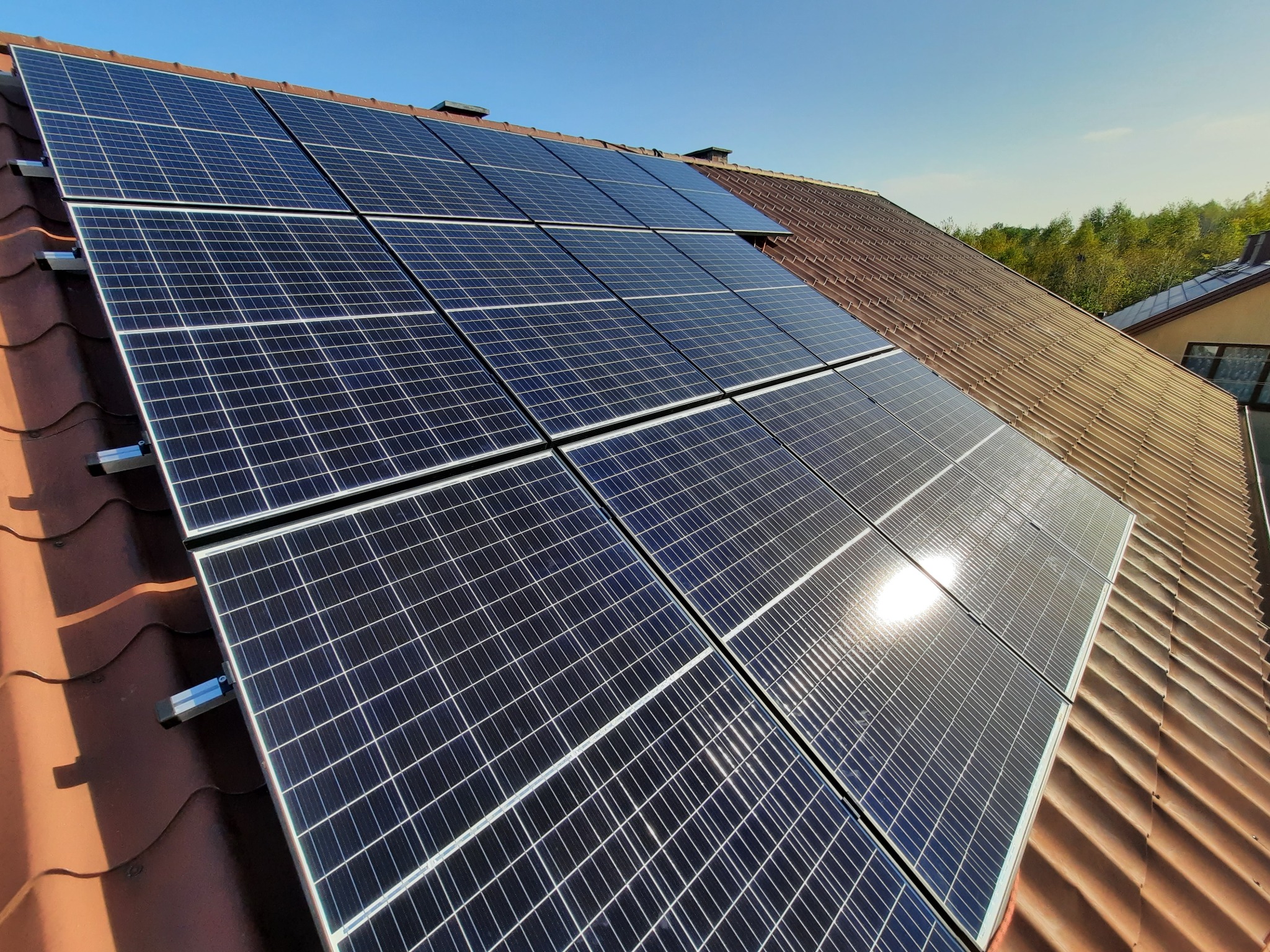 Panele fotowoltaiczne zamontowane na brązowym dachu z dachówki ceramicznej, widoczne mocowania paneli, błękitne niebo w tle.