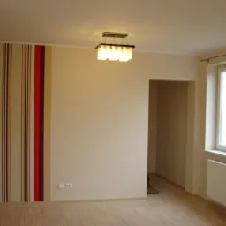 Pusty pokój po remoncie: beżowe ściany, panel z tapetą w pionowe pasy (czerwony, beżowy, brązowy), okno z białymi ramami i grzejnikiem, drewniana podłoga, sufit z lampą.