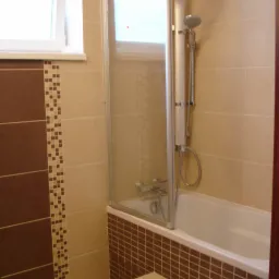 Wykończona łazienka z wanną obudowaną brązową mozaiką, szklany parawan nawannowy, beżowe płytki ścienne z dekoracyjnym paskiem z mozaiki, białe okno.