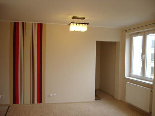 Pusty pokój po remoncie: beżowe ściany, panel z tapetą w pionowe pasy (czerwony, beżowy, brązowy), okno z białymi ramami i grzejnikiem, drewniana podłoga, sufit z lampą.