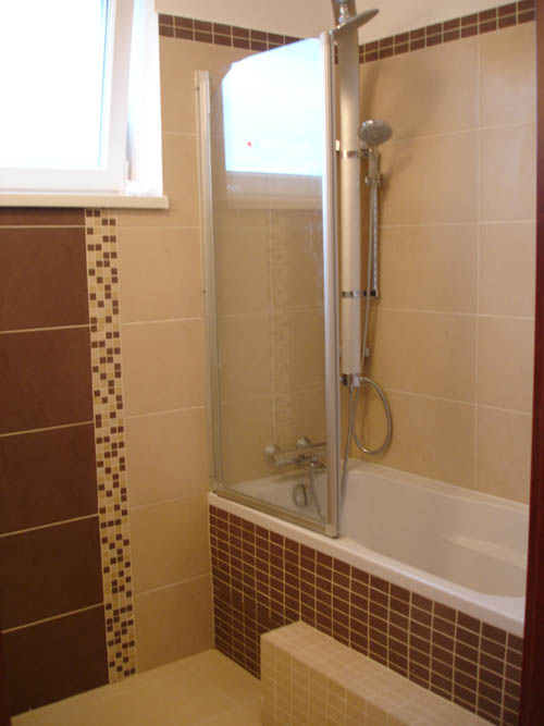 Wykończona łazienka z wanną obudowaną brązową mozaiką, szklany parawan nawannowy, beżowe płytki ścienne z dekoracyjnym paskiem z mozaiki, białe okno.