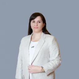 Portret kobiety w beżowym żakiecie i białej koszulce z subtelnym wzorem, pozującej na jednolitym szarym tle, z delikatnym uśmiechem i złotym zegarkiem na nadgarstku.