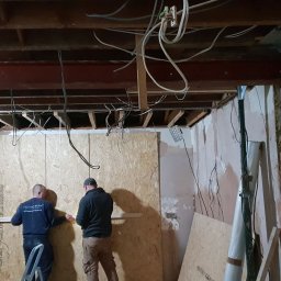 Sebko - Dwóch mężczyzn na drabinie montuje płytę OSB na ścianie w pomieszczeniu w trakcie remontu, widoczne odsłonięte kable, wsporniki sufitowe i fragmenty starego tynku.