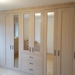 Sebko - Duża, jasna szafa wnękowa z lustrami, z drewnianymi frontami i metalowymi uchwytami, zamontowana w pokoju z drewnianą podłogą i białymi ścianami.