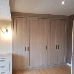 Sebko - Duża, jasna szafa wnękowa z sześcioma drzwiami i podłużnymi uchwytami, dopasowana do skosu sufitu na poddaszu, z komodą z szufladami po lewej stronie i otwartymi drzwiami po prawej.