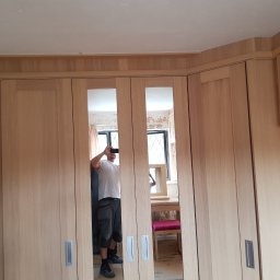 Sebko - Wbudowana szafa z jasnego drewna z lustrzanymi drzwiami, widoczne niedokończone ściany i odbicie osoby robiącej zdjęcie telefonem.