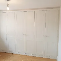 Biała, zabudowana szafa wnękowa z sześcioma drzwiami i okrągłymi, szklanymi gałkami, zamontowana w pokoju w Łodzi z drewnianą podłogą i widoczną żarówką na suficie.