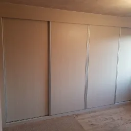 Beżowa szafa wnękowa z przesuwnymi drzwiami, z drewnianym wykończeniem, zamontowana w pokoju z surowymi ścianami i drewnianą podłogą.