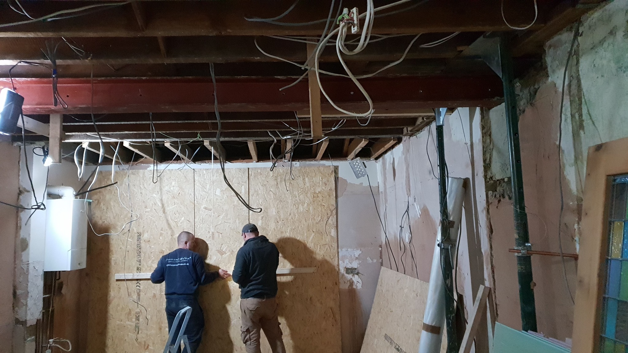 Dwóch mężczyzn na drabinie montuje płytę OSB na ścianie w pomieszczeniu w trakcie remontu, widoczne odsłonięte kable, wsporniki sufitowe i fragmenty starego tynku.