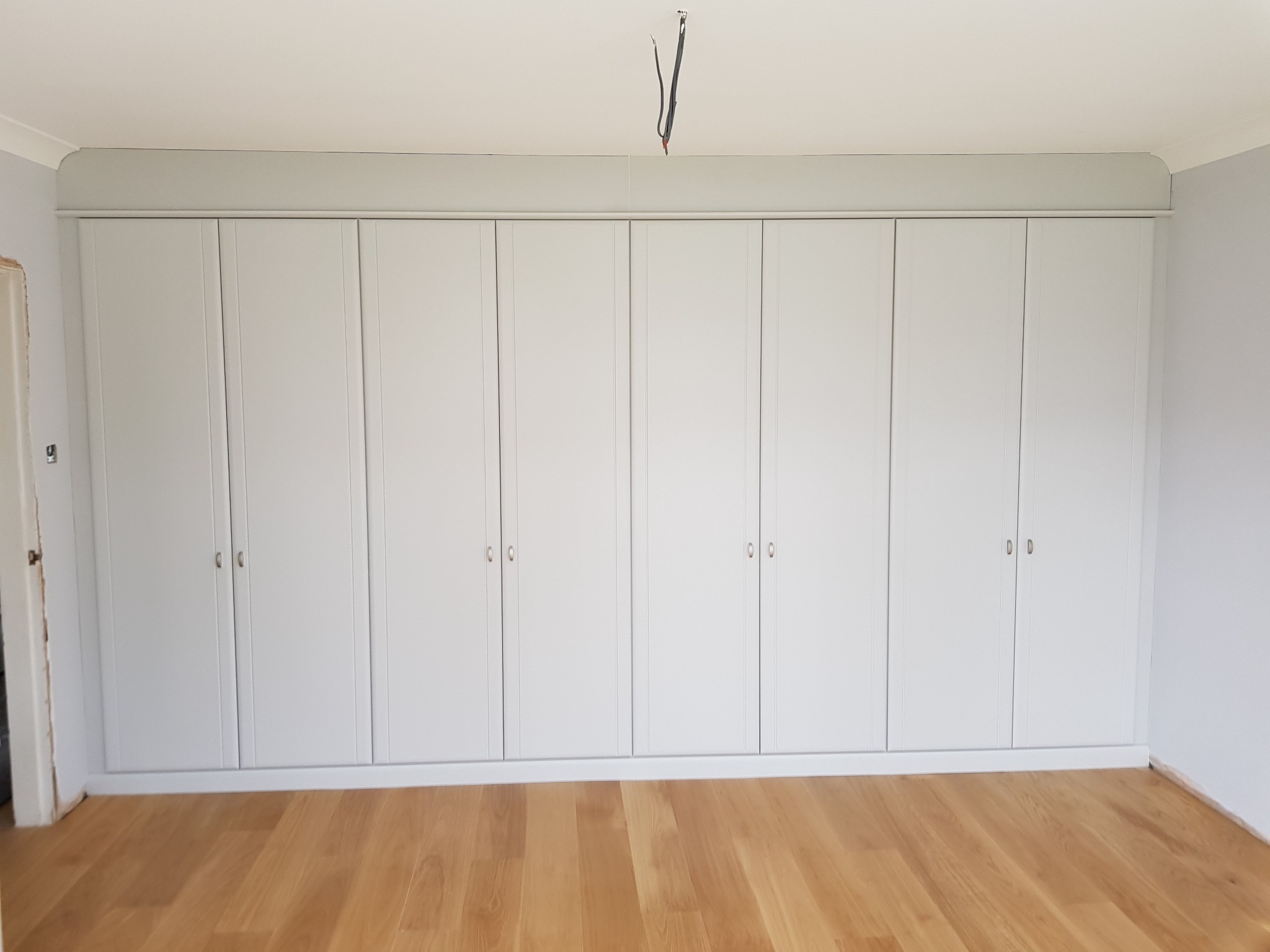 Wbudowana szafa z jasnoszarymi frontami drzwiowymi, dopasowana do koloru ścian, z drewnianą podłogą w nowym mieszkaniu; widoczne wystające przewody elektryczne z sufitu.