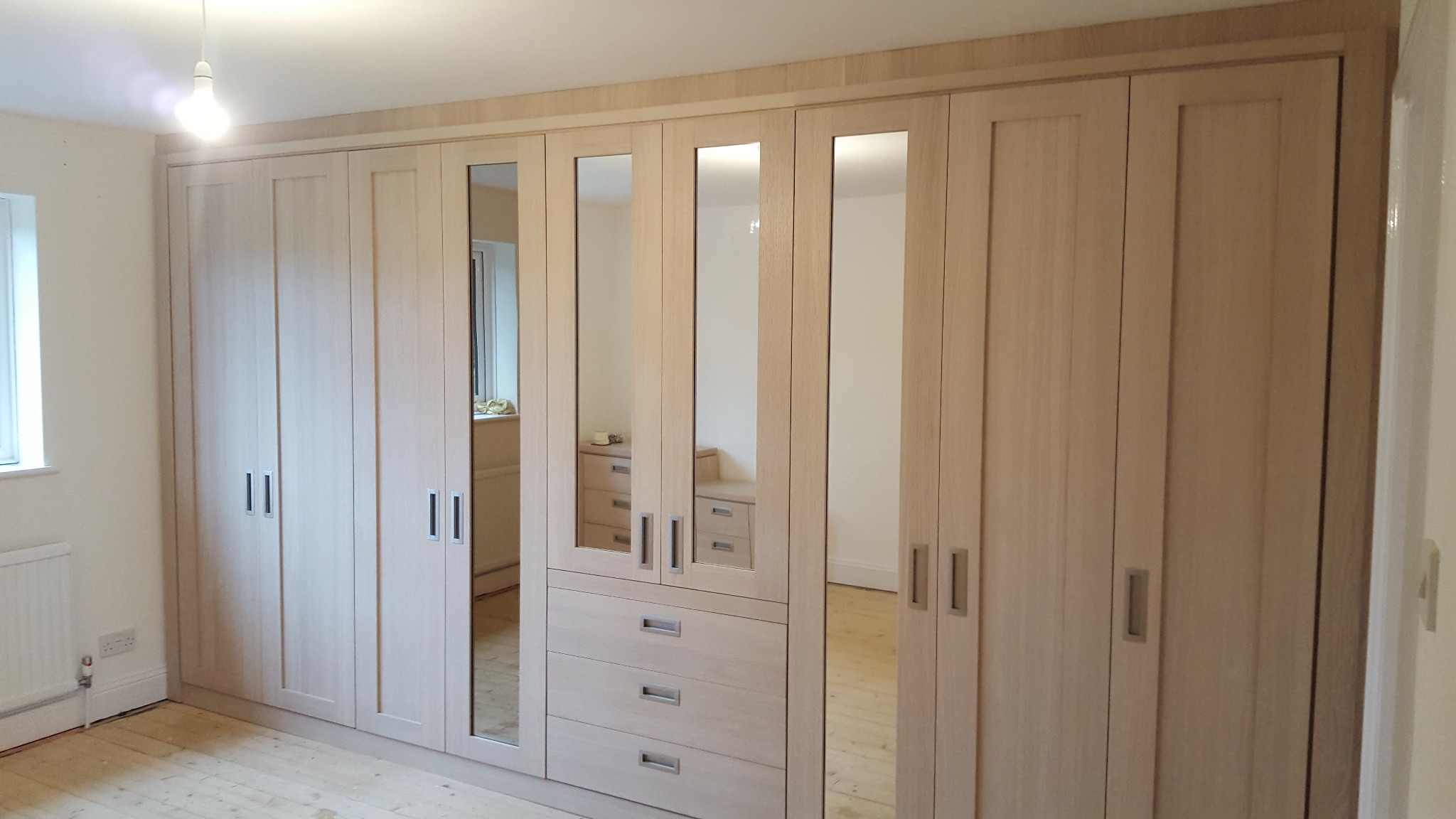 Duża, jasna szafa wnękowa z lustrami, z drewnianymi frontami i metalowymi uchwytami, zamontowana w pokoju z drewnianą podłogą i białymi ścianami.