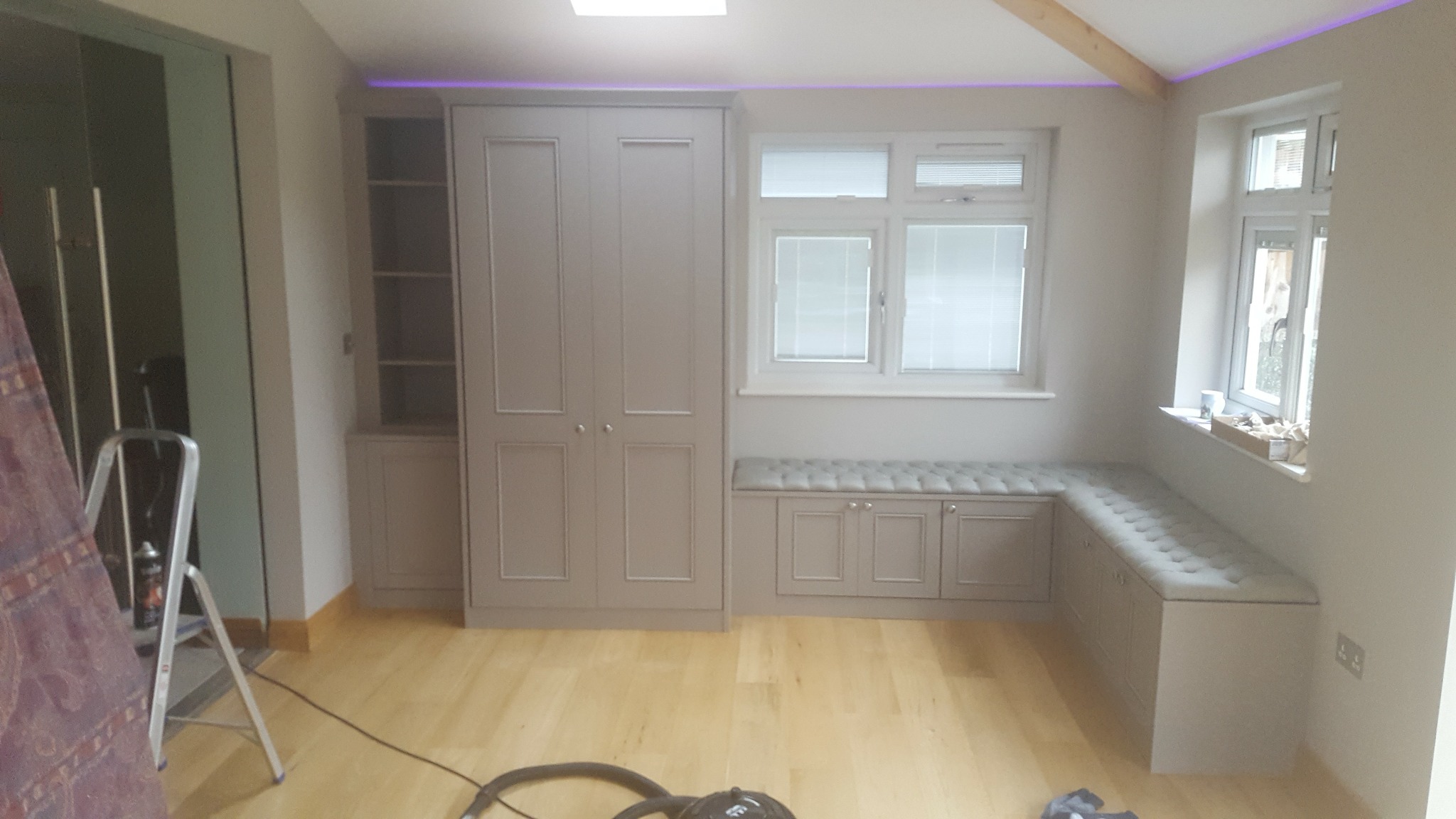 Szara zabudowa meblowa w pokoju z drewnianą podłogą, obejmująca szafę, regał i siedziska pod oknami z tapicerowanymi poduszkami, widoczny fragment drabiny i odkurzacza.