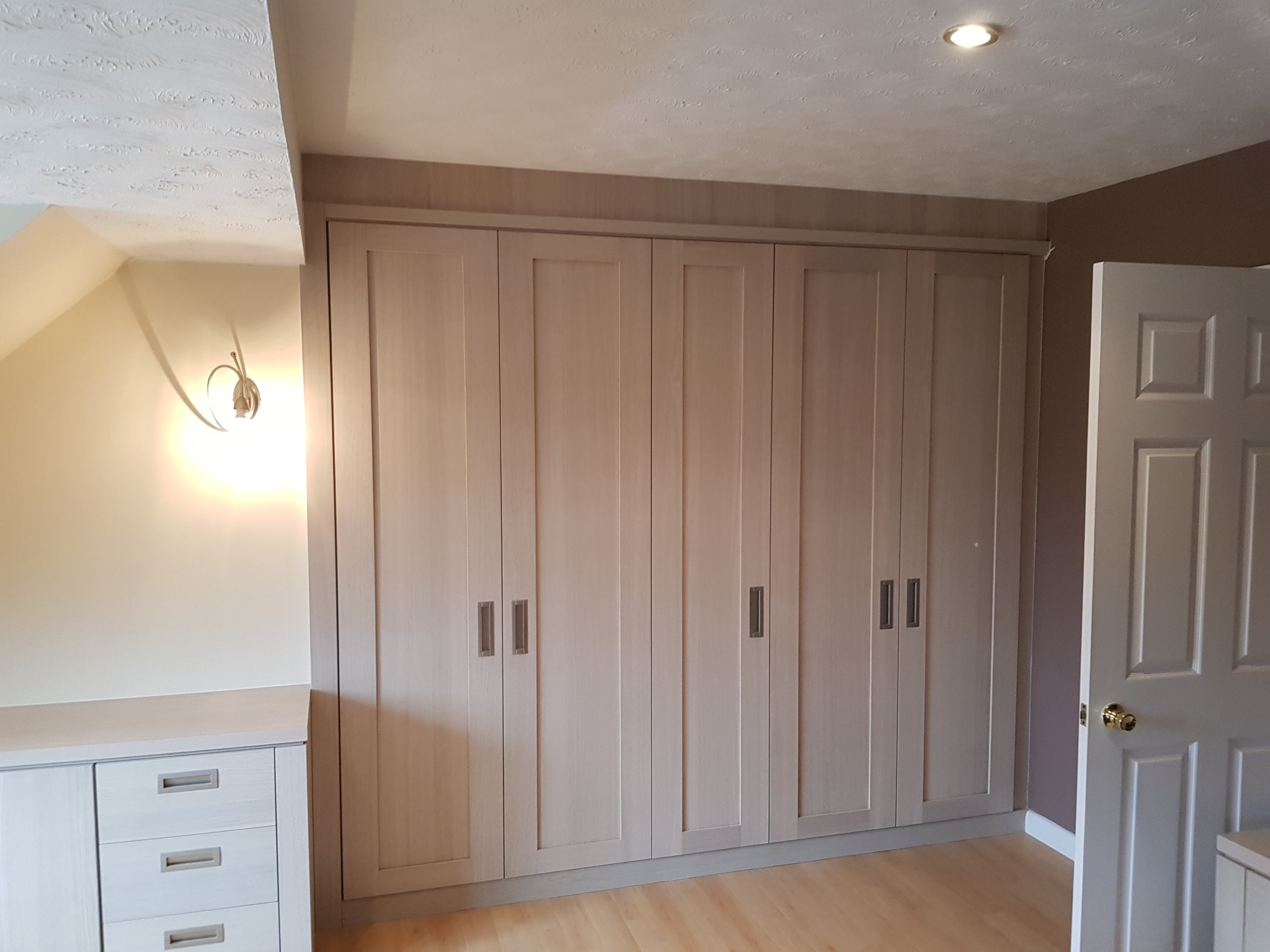 Duża, jasna szafa wnękowa z sześcioma drzwiami i podłużnymi uchwytami, dopasowana do skosu sufitu na poddaszu, z komodą z szufladami po lewej stronie i otwartymi drzwiami po prawej.