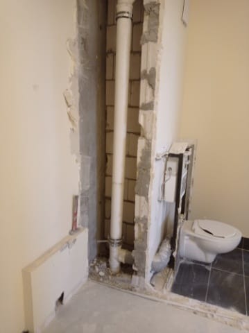 Fragment ściany z odsłoniętą instalacją kanalizacyjną w trakcie remontu łazienki, widoczny fragment toalety i czarne płytki podłogowe.