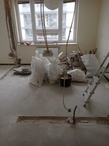 Wnętrze w trakcie wyburzania ścian z widocznymi workami z gruzem, odsłoniętymi przewodami i bruzdą w podłodze, drabina oparta o ścianę, duże okno w tle.
