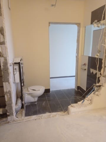 Wnętrze łazienki w trakcie demontażu: widoczne częściowo usunięte płytki, odsłonięte instalacje i toaleta. Ślady po skuciu ścian i podłóg.