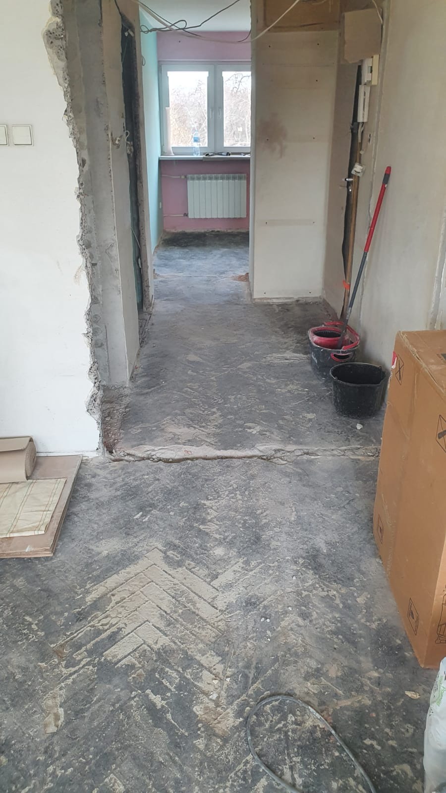 Wnętrze w trakcie remontu: usunięte ściany, odsłonięta podłoga z wzorem jodełki, widoczne ślady po pracach, wiadro i mop w tle, kartonowe pudło z prawej strony.