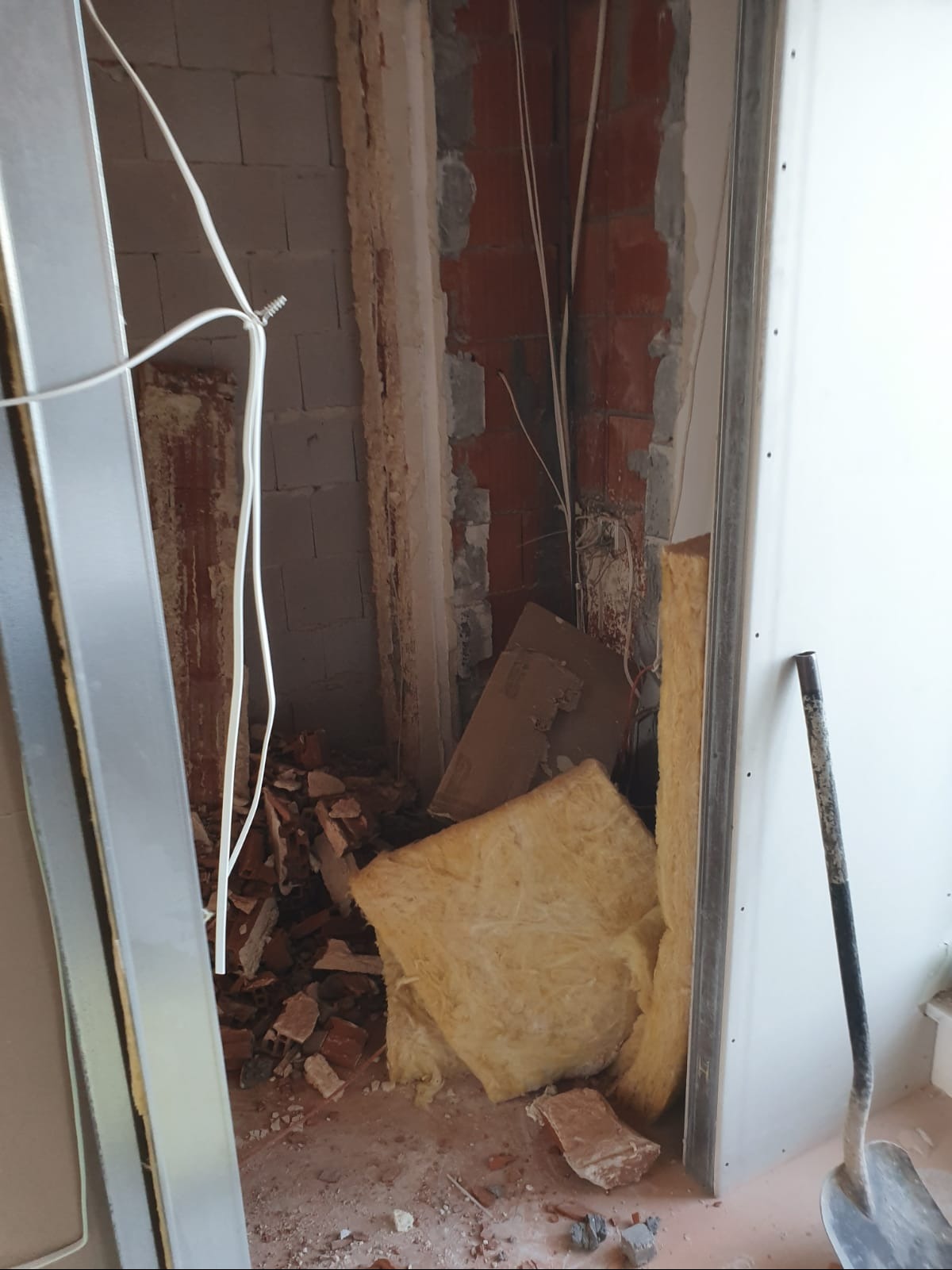 Wnętrze w trakcie demontażu ściany z widocznymi fragmentami cegieł, przewodami elektrycznymi, wełną mineralną i łopatą.