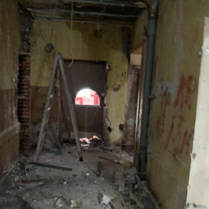 Wnętrze w trakcie wyburzania, widoczne odsłonięte cegły, stara drabina oparta o ścianę, gruz na podłodze i fragmenty instalacji elektrycznej.