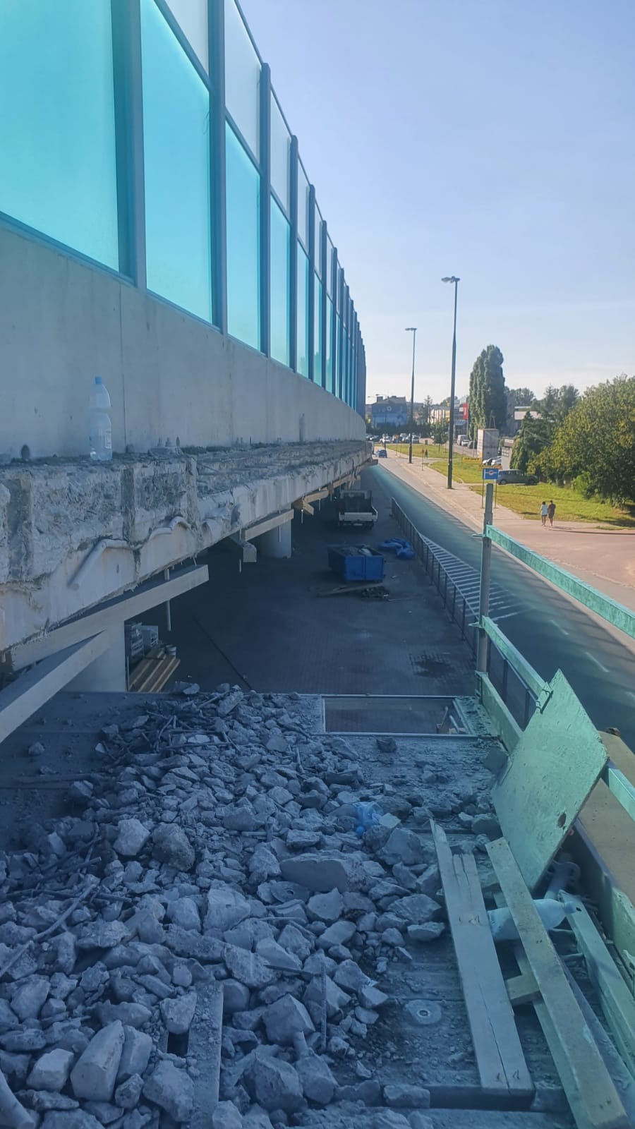 Demontaż fragmentu betonowej konstrukcji mostu lub wiaduktu, widoczne gruz i pozostałości po rozbiórce, w tle ulica i budynki.