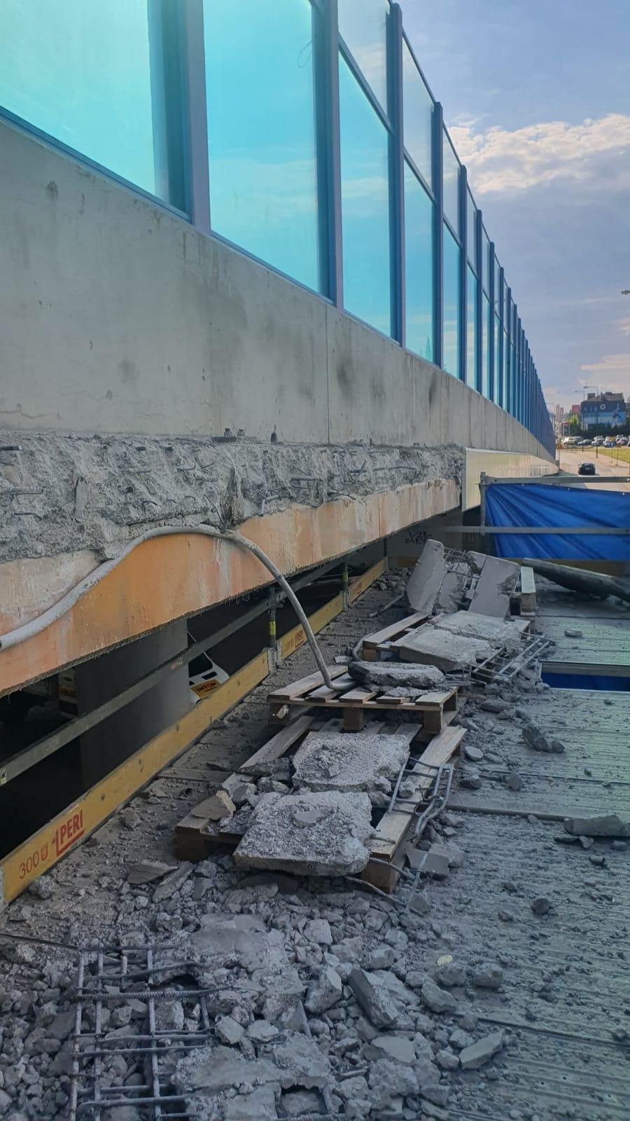 Fragment zdemontowanego betonowego wiaduktu z widocznym zbrojeniem, pozostałościami betonu i deskowaniem, na tle błękitnej bariery dźwiękochłonnej.