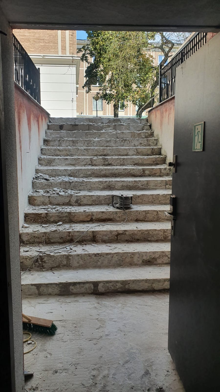 Betonowe schody w trakcie demontażu, widoczne gruz i narzędzia, otwarte drzwi z symbolem ewakuacyjnym, budynek w tle.