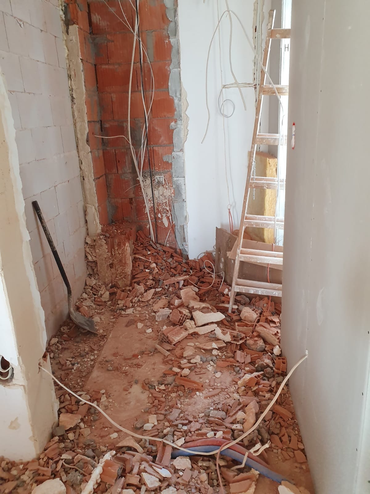 Wnętrze w trakcie prac rozbiórkowych: gruz z cegieł, szpachla na ścianach, wystające kable, drabina oparta o ścianę, widoczna łopata.