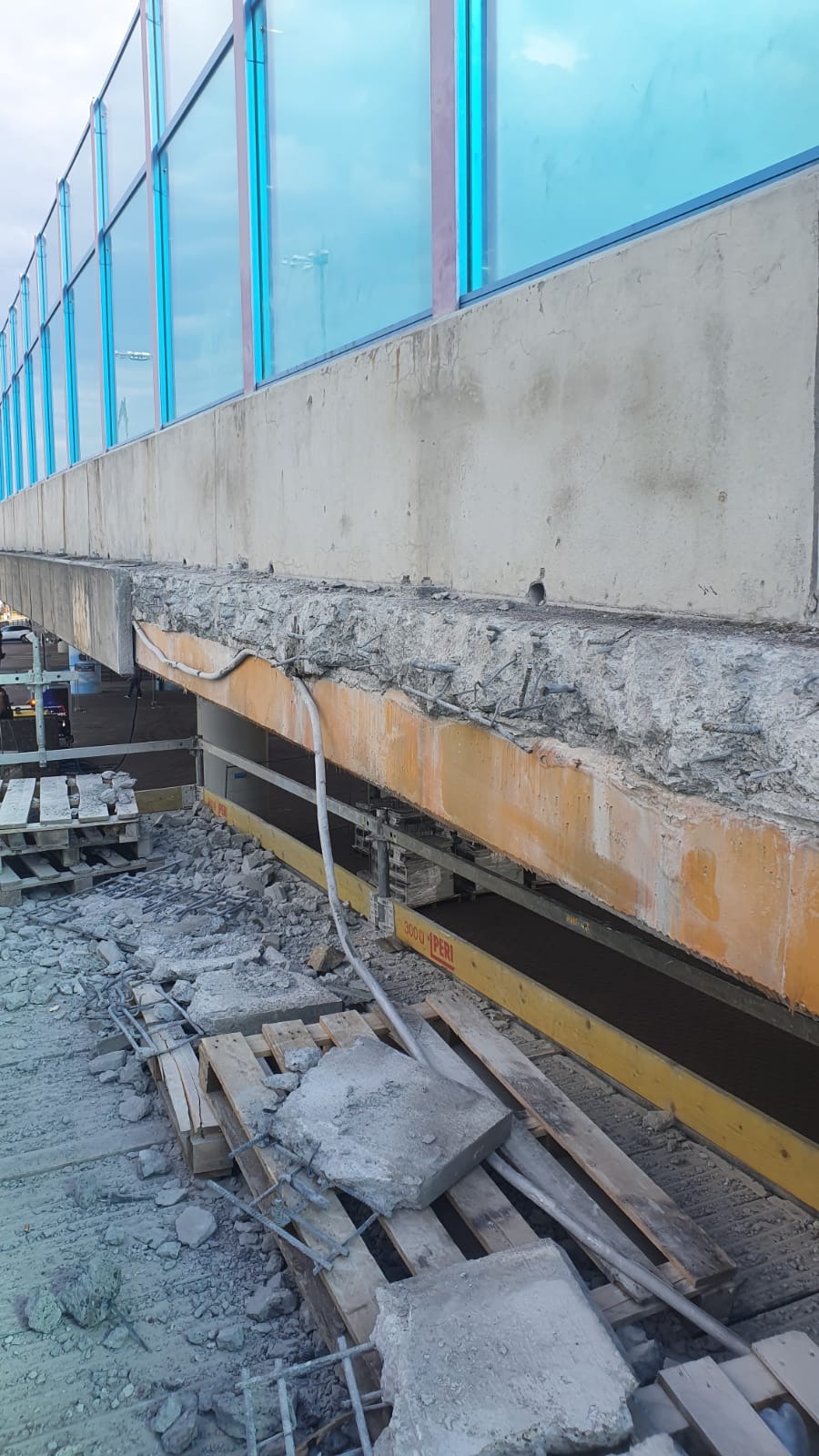 Uszkodzony betonowy element konstrukcyjny z odsłoniętym zbrojeniem, podparty żółtą konstrukcją wzmacniającą, gruz betonowy na ziemi, w tle błękitne panele.