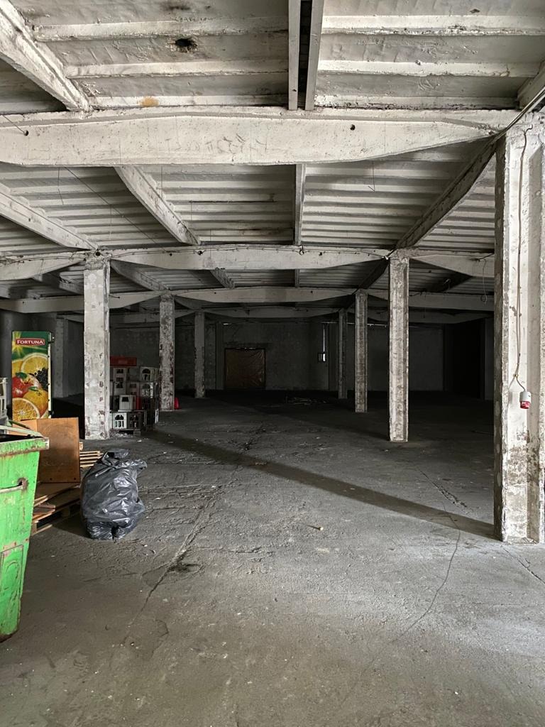 Pusta hala przemysłowa ze starymi betonowymi kolumnami i stropem żebrowym, widoczny śmietnik i automat z napojami w tle, podłoga betonowa z widocznymi pęknięciami.