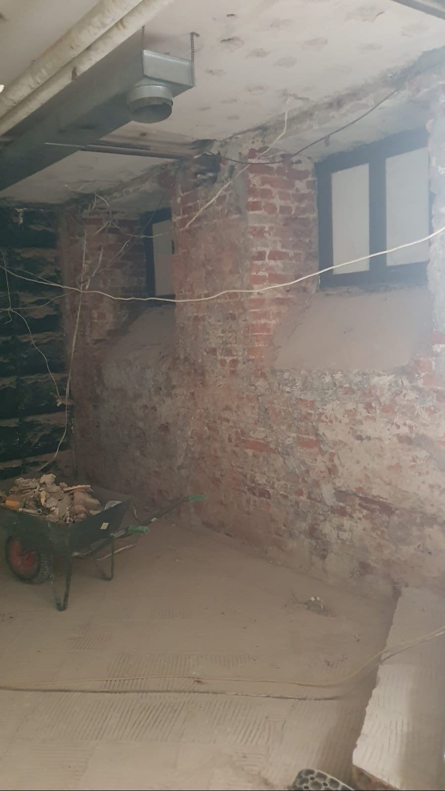 Wnętrze w trakcie prac wyburzeniowych: odsłonięta cegła, częściowo otynkowane ściany, gruz w taczce, widoczne przewody i instalacje pod sufitem.