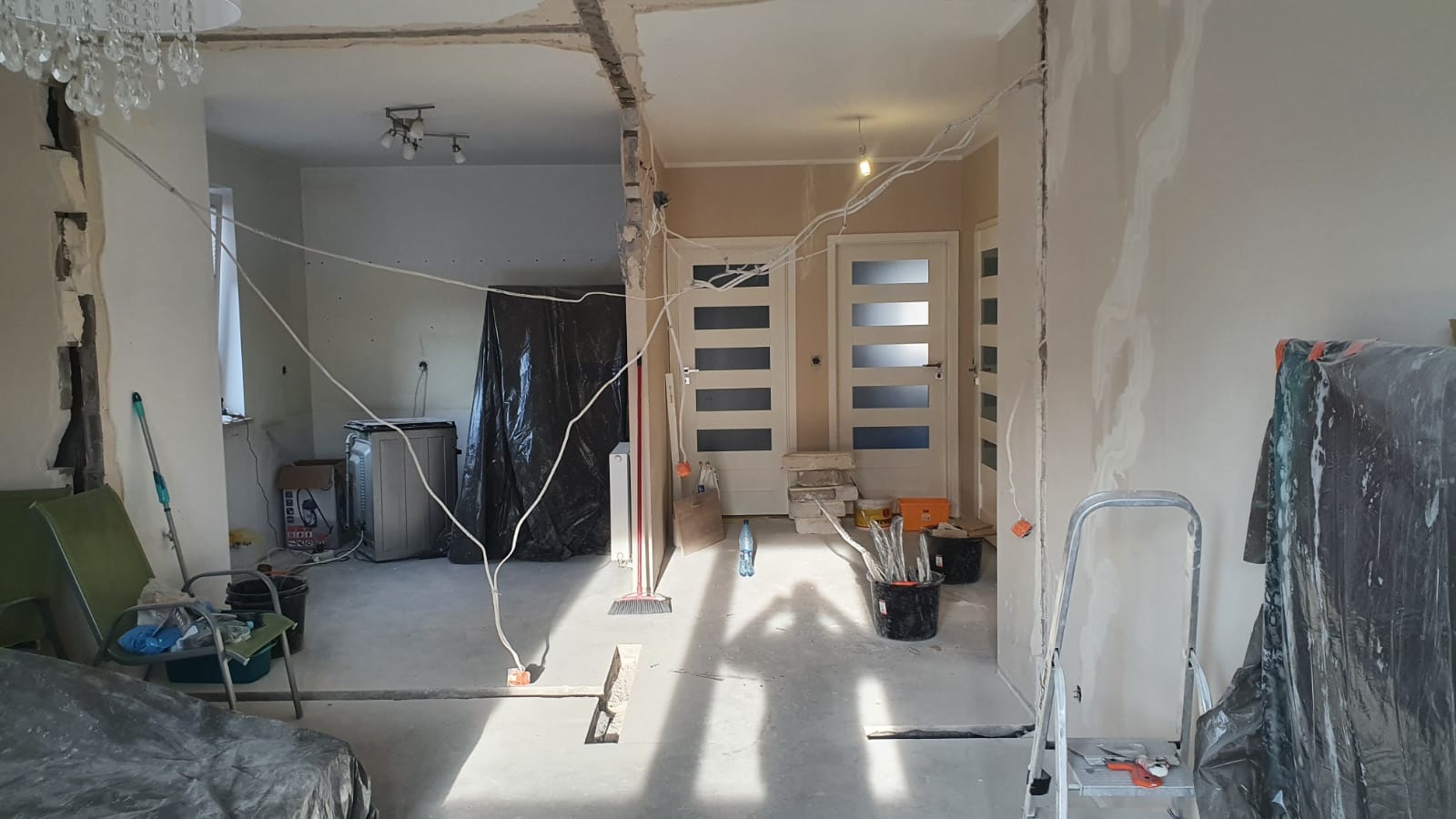 Wnętrze w trakcie wyburzania ściany działowej, widoczne przewody elektryczne, zabezpieczone meble, ślady po kuciu na ścianach, przygotowane narzędzia i materiały.