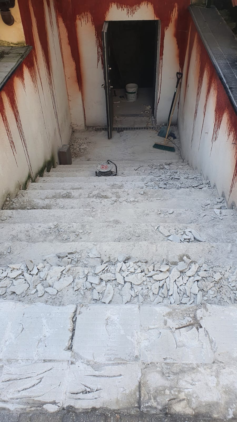 Schody betonowe w trakcie demontażu, z widocznymi odłamkami gruzu, ślady czerwonej farby na ścianach oraz otwarte drzwi do piwnicy z narzędziami w tle.
