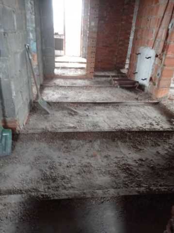 Wnętrze budynku w trakcie prac przygotowawczych do wylewki, widoczne szalunki, ziemia i ceglane ściany w surowym stanie, z widoczną łopatą opartą o ścianę.