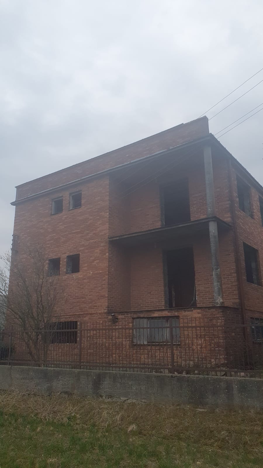 Dwupiętrowy budynek z czerwonej cegły w stanie surowym zamkniętym, z widocznymi otworami okiennymi i balkonami, otoczony metalowym ogrodzeniem i skąpą roślinnością na pochmurnym tle.