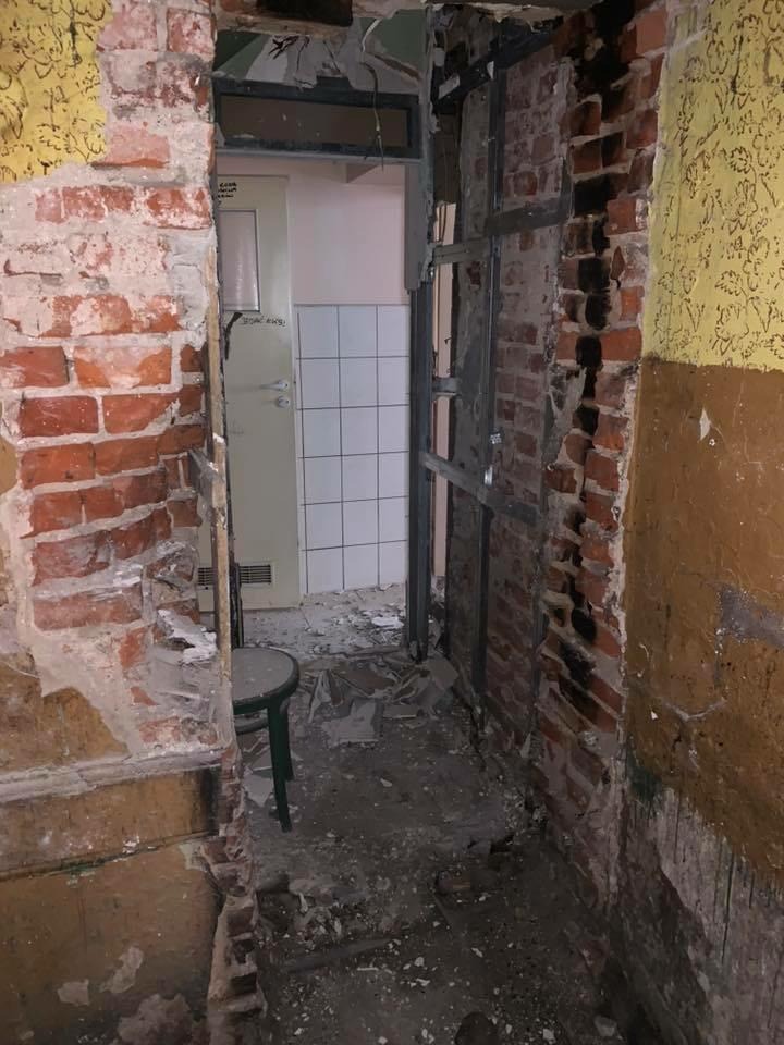 Wnętrze w trakcie demontażu: odsłonięta cegła, fragmenty tapety, metalowa konstrukcja pod płytę gipsową i widok na pomieszczenie z białymi płytkami. Widoczny bałagan i pozostałości po pracach.