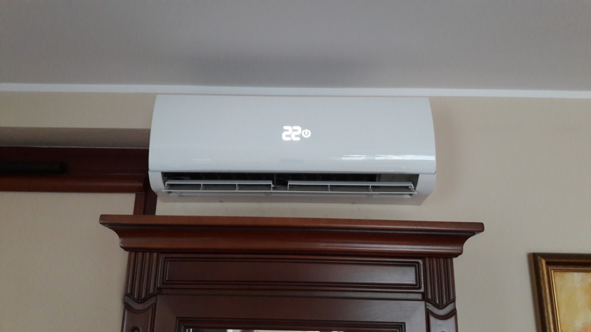 Biały klimatyzator ścienny z wyświetlaczem temperatury 22 stopnie Celsjusza, zamontowany nad ciemnobrązową komodą w pokoju.