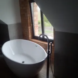 Wolnostojąca, owalna wanna z chromowaną baterią podłogową, ustawiona przy oknie w łazience na poddaszu z ceglanym filarem.