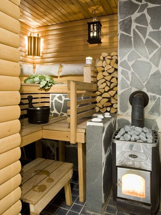 Wnętrze drewnianej kabiny z piecem opalanym drewnem, kamienną ścianą, drewnianymi ławkami z poduszkami i śladami stóp na taborecie.