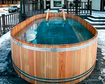 Drewniana beczka kąpielowa z drabinką, wypełniona wodą, otoczona śniegiem na drewnianym tarasie. W tle widoczny budynek i zaparkowany samochód.