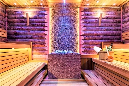 Wnętrze sauny wyłożone drewnem z podświetleniem, z kamiennym piecem centralnym i akcesoriami do kąpieli. Widoczne drewniane ławy i oświetlenie punktowe.